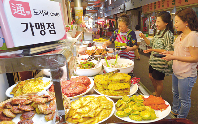 Two Tongin Community employees demonstrate how to use Dosirak Cafe Tong at one of the cafe’s affiliate stores. After purchasing golden tokens, customers can use them to buy banchan (side dishes) and other eats at affiliate stores in Tongin Market.