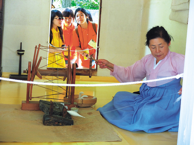 A group of students look in on a woman dressed in traditional clothes as she works a spindle at the Korean Folk Village in Suwon. (Maryann Wright/The Korea Herald)