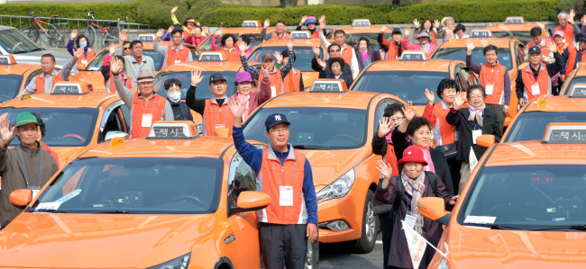 Taxi drivers and senior citizens wave for a photo in the parking lot of Guro-gu Office in Seoul on Tuesday before departing for tourist attractions on Ganghwado Island. The cabbies gave old or disabled people in the district a complimentary ride to tourist sites as a voluntary service.(Kim Myung-sub/The Korea Herald)
