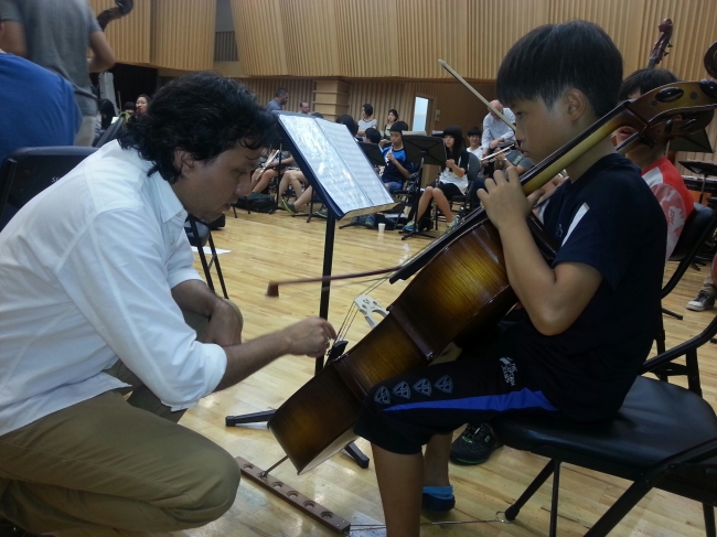 Venezuelan cellist Aristides Rivas (left) helps a Korean child hone his craft during rehearsal in an El Sistemainspiredintensive 10-day music camp at the Sejong Center for the Performing Arts in Seoul on Friday. (Venezuela Embassy)