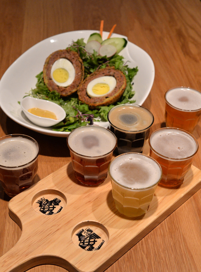 7Brau Pub’s beer sampler (front) features IPA, Mild Ale, Pilsner, Fake Lager, Kristal Weizen and Stout. The Gangnam outpost’s take on the classic Scotch egg (back) pairs well with the caramel notes of the Fake Lager.(Lee Sang-sub/The Korea Herald)