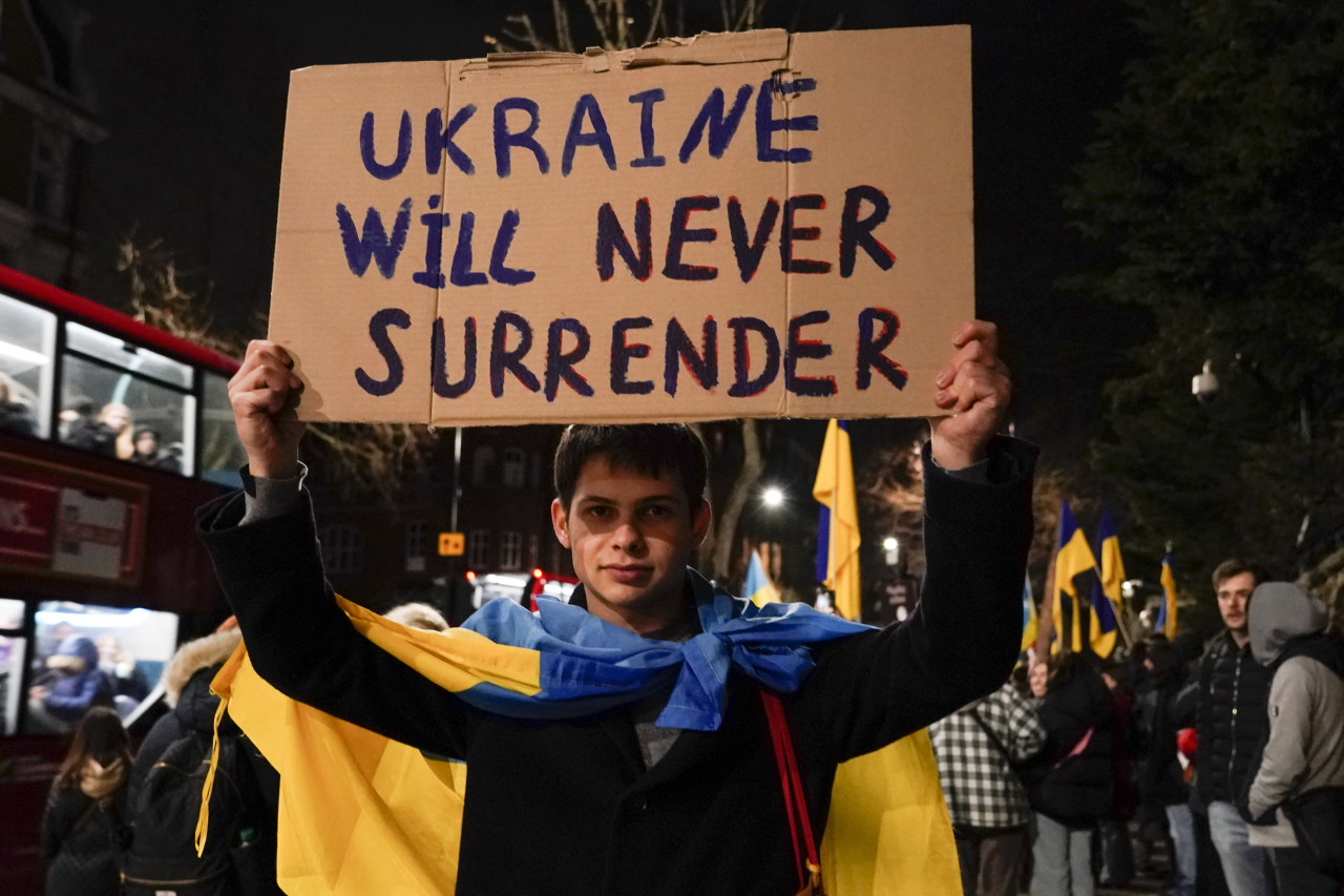 A man wearing a Ukrainian flag in front of the Russian Embassy in London holds a sign with the phrase “Ukraine will never surrender” on Wednesday. (Yonhap)