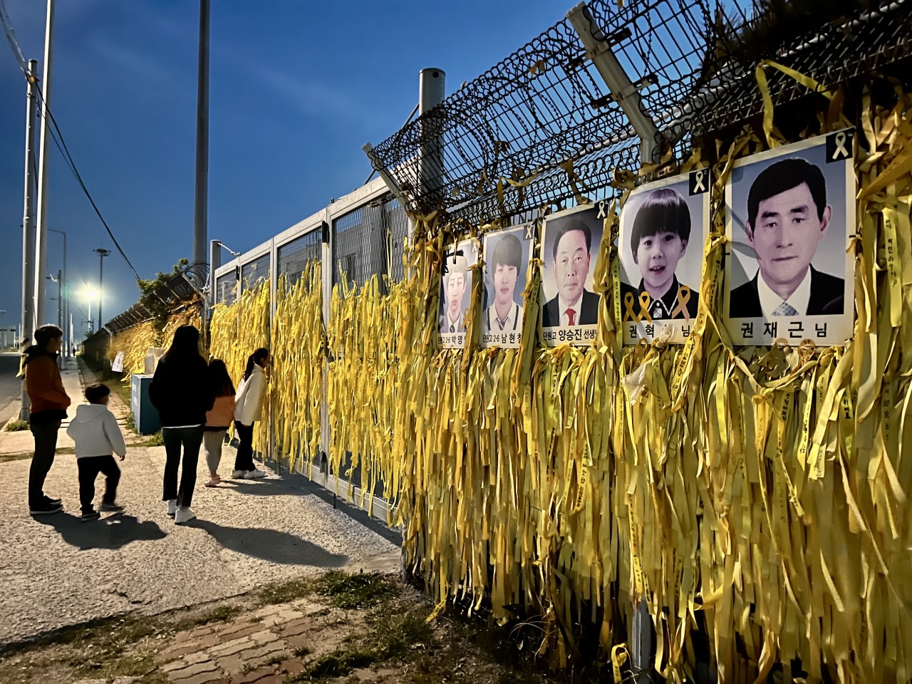 On a chain-link fence, yellow ribbons of tribute for victims and pictures of the missing have been hung. The recovered ferry lays on the other side of the wall. (Kim Arin/The Korea Herald)