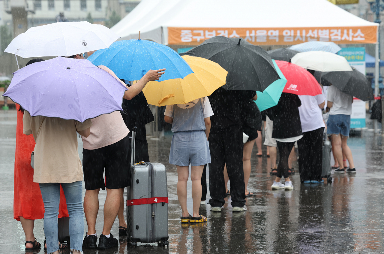 People carrying umbrellas stand in line for COVID-19 tests at a makeshift testing station at Seoul Plaza in central Seoul on Sunday, as the country has experienced a new virus wave. (Yonhap)