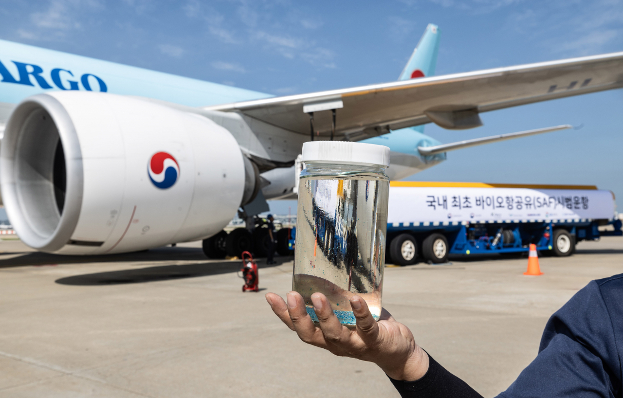 A Korean Air representative holds a bottle of Neste's Sustainable Aviation Fuel (SAF), with the refueled aircraft in the background. Aviation fuel is clear because the refining process removes impurities to meet strict safety and performance specifications for aircraft engines. (Korean Air)