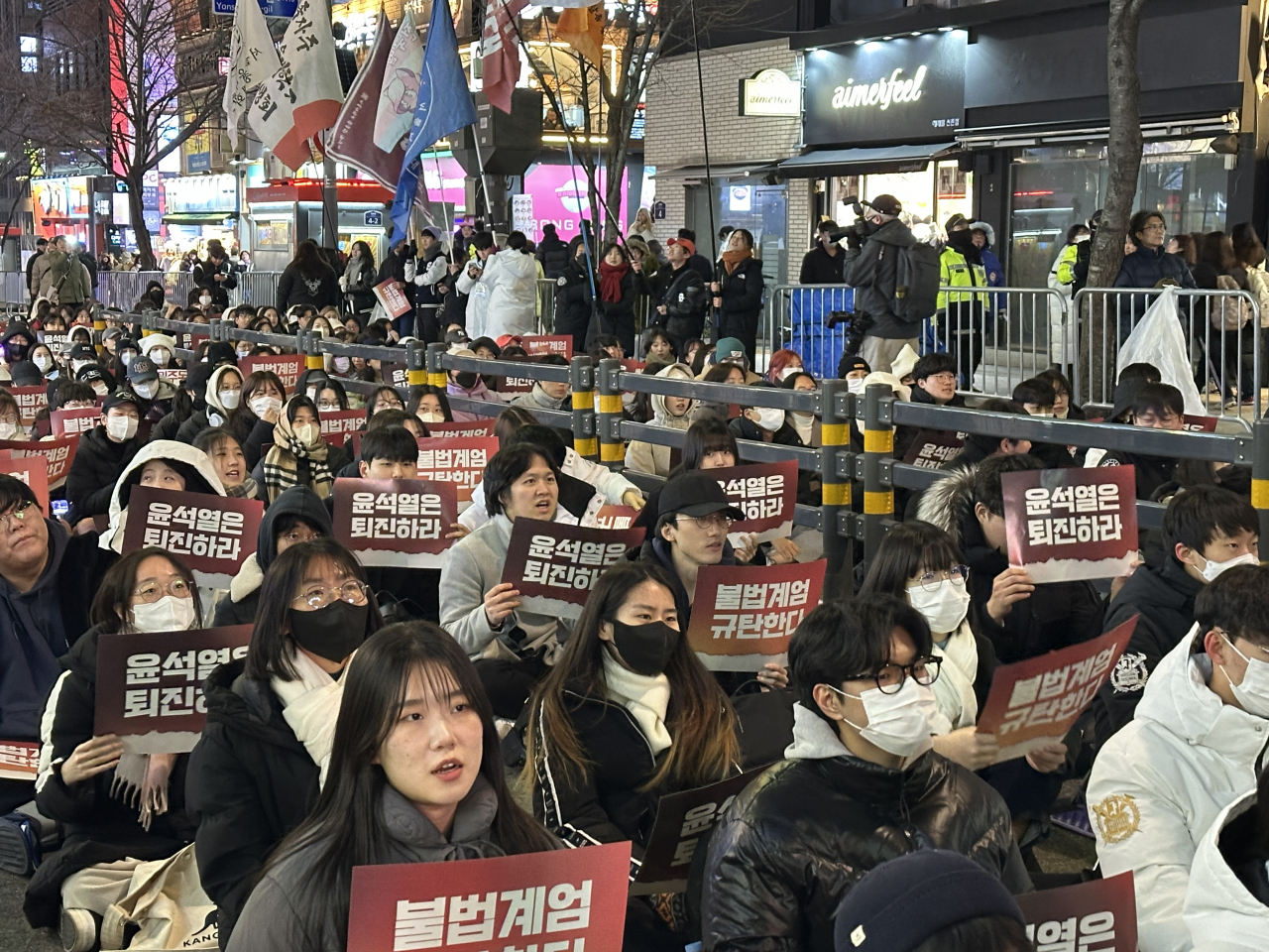 University students sit in the streets of Sinchon in Seodaemun-gu, western Seoul, holding signs that read “We condemn martial law” and “Yoon Suk Yeol, step down immediately” during a mass rally led by university students on Friday. (Lee Jung-joo/The Korea Herald)