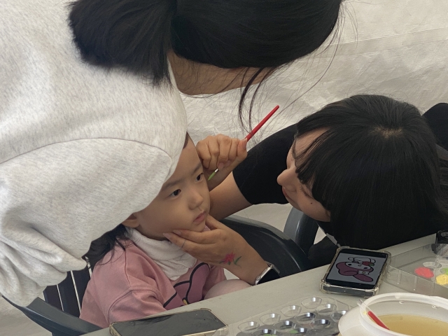 An art student from Kaywon University offers face painting to a young visitor at the Wellness Seoul 2024 booth, Sunday. (Hwang Joo-young/The Korea Herald)