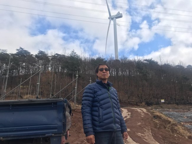 Local farmer Park Chung-lak, who grows apples in the Honggye-ri village in North Gyeongsang Province, speaks in front of his farm. Behind Park lies a 100-meter-high wind turbine that was installed as part of Korea’s renewable energy policies. Bak Se-hwan/The Korea Herald