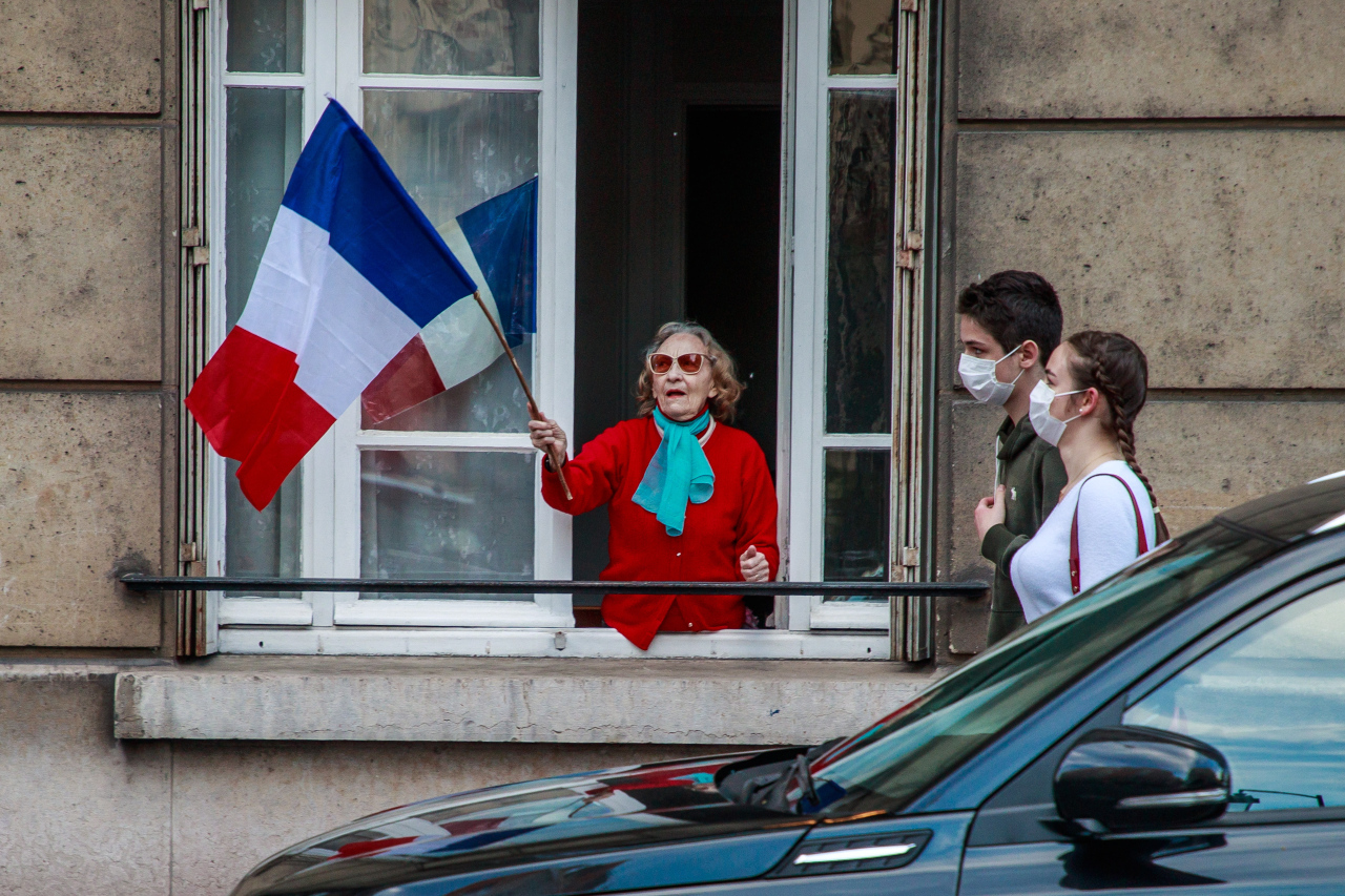 A couple wearing protectives face masks pass by a building where a woman holds a French flags as the neighbors applaud every evening at 8pm from their windows to support French medical staff amid the ongoing coronavirus COVID-19 pandemic in Saint Mande, near Paris, France, 02 May 2020. (AFP-Yonhap)